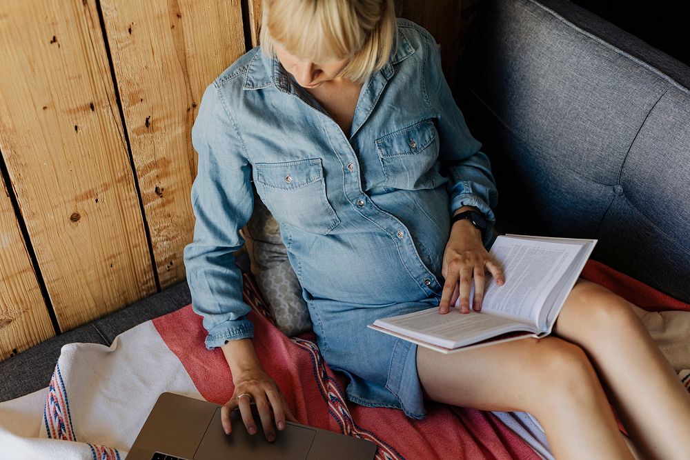 Pregnant woman searching on a website from her laptop while reading a book on a couch