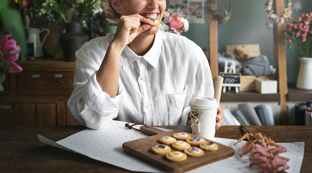 Smiling woman enjoying homemade cookies with floral garnish and a cup of coffee in a flower shop