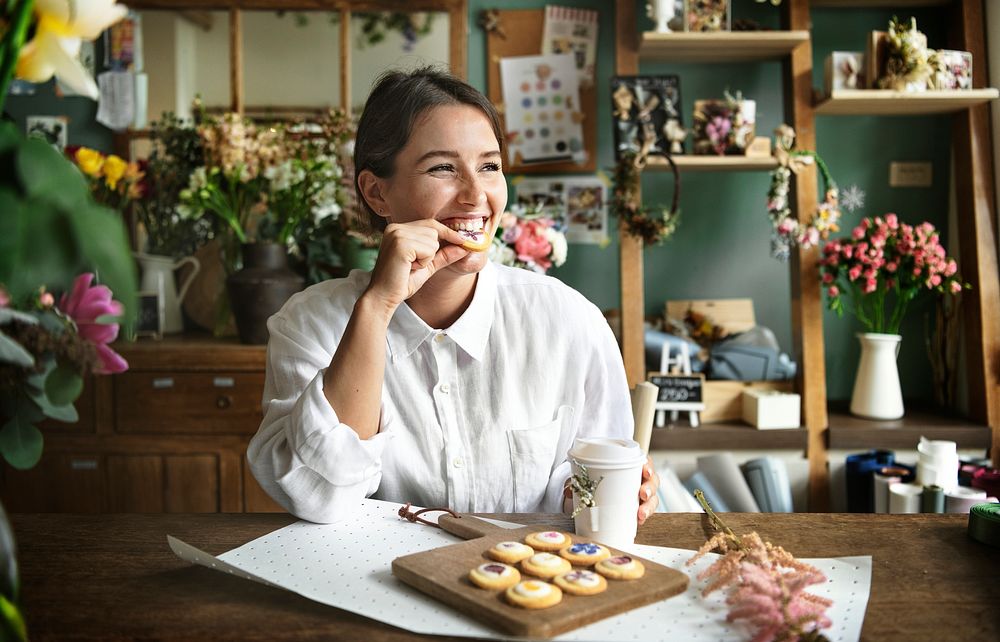 Smiling woman enjoying homemade cookies with floral garnish and a cup of coffee in a flower shop