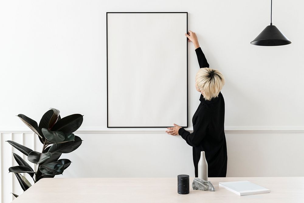 Blond haired Asian woman hanging a blank frame on a white wall