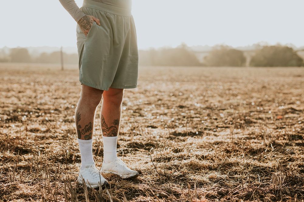 Tattooed man in running shorts standing in countryside at sunset