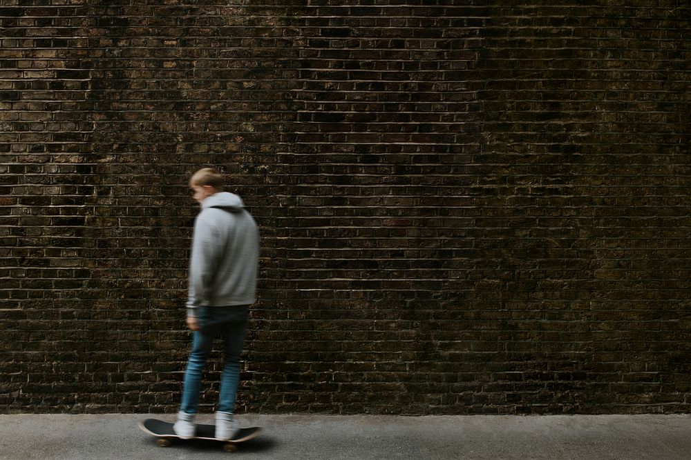 Man in gray hoodie skating in city, passing grunge brick wall