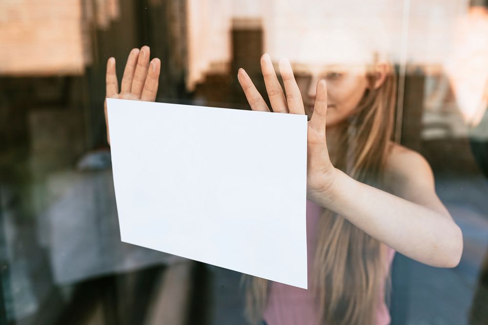 Blond girl showing blank paper sign through glass window, the new normal