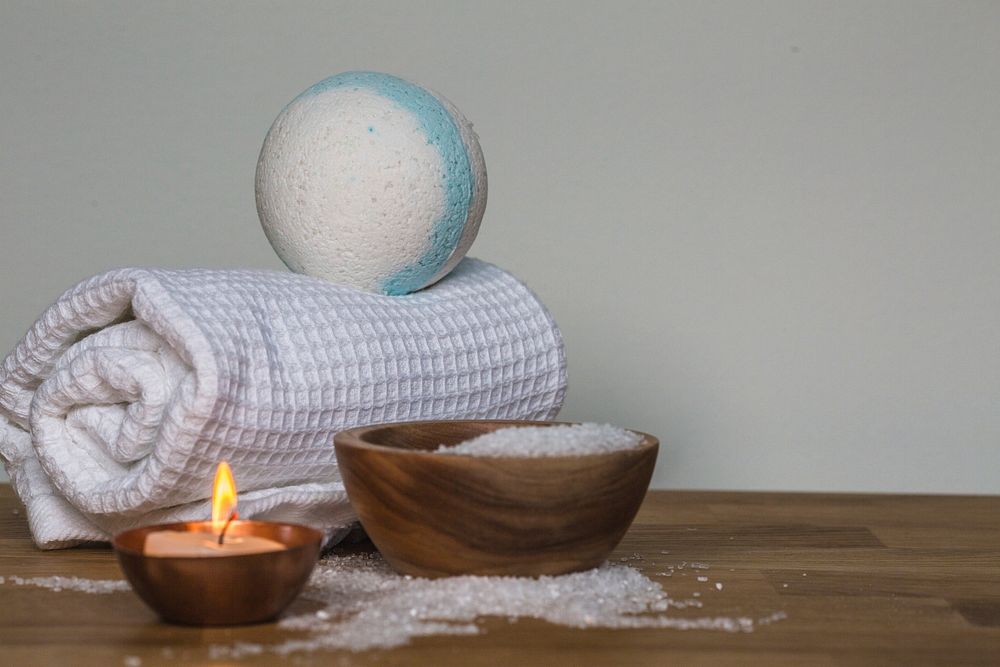 Blue and white luxury bath bomb on white towel, next to wooden bowl full of epsom salts and lit candle.