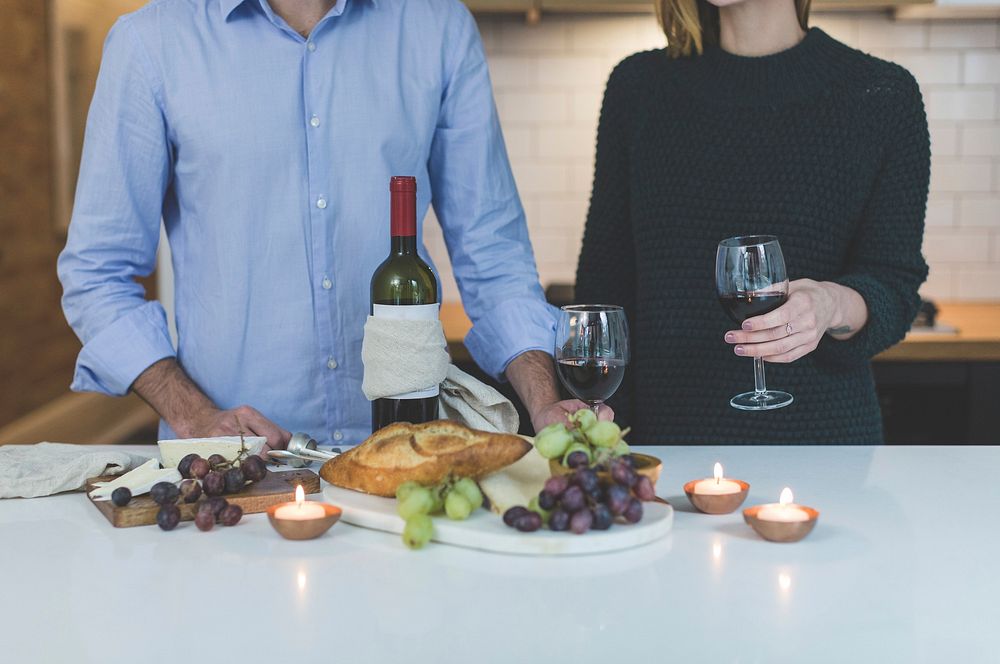 Free man and woman standing behind wine and appetizer spread at kitchen counter, public domain CC0 photo.