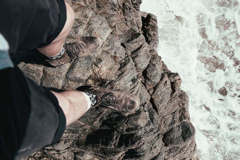 A shot from above of a bare-legged man in beat up climbing boots standing on the edge of a cliff overlooking rushing waters…