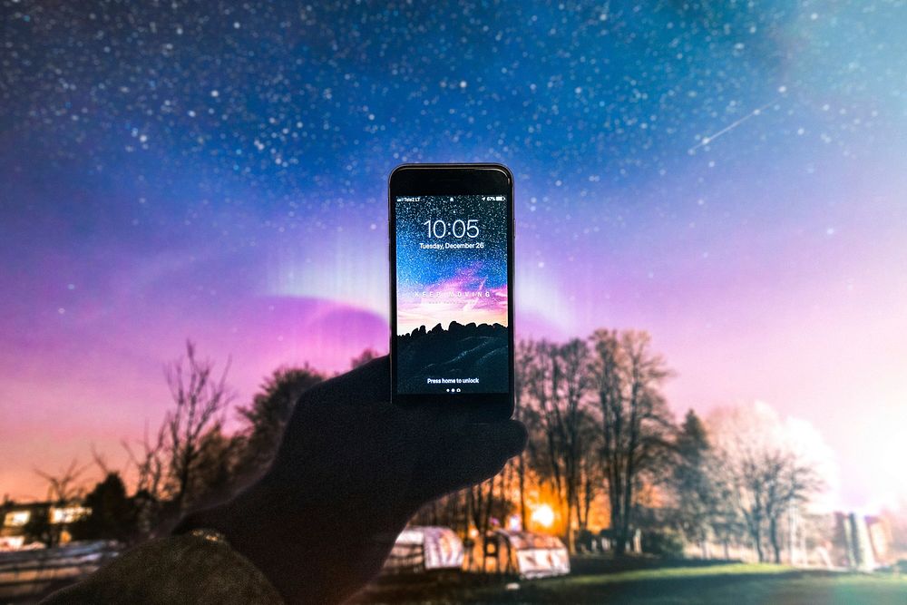 A hand holds up an iPhone against a country star-filled sky with the sun setting. The iPhone wallpaper has an almost…