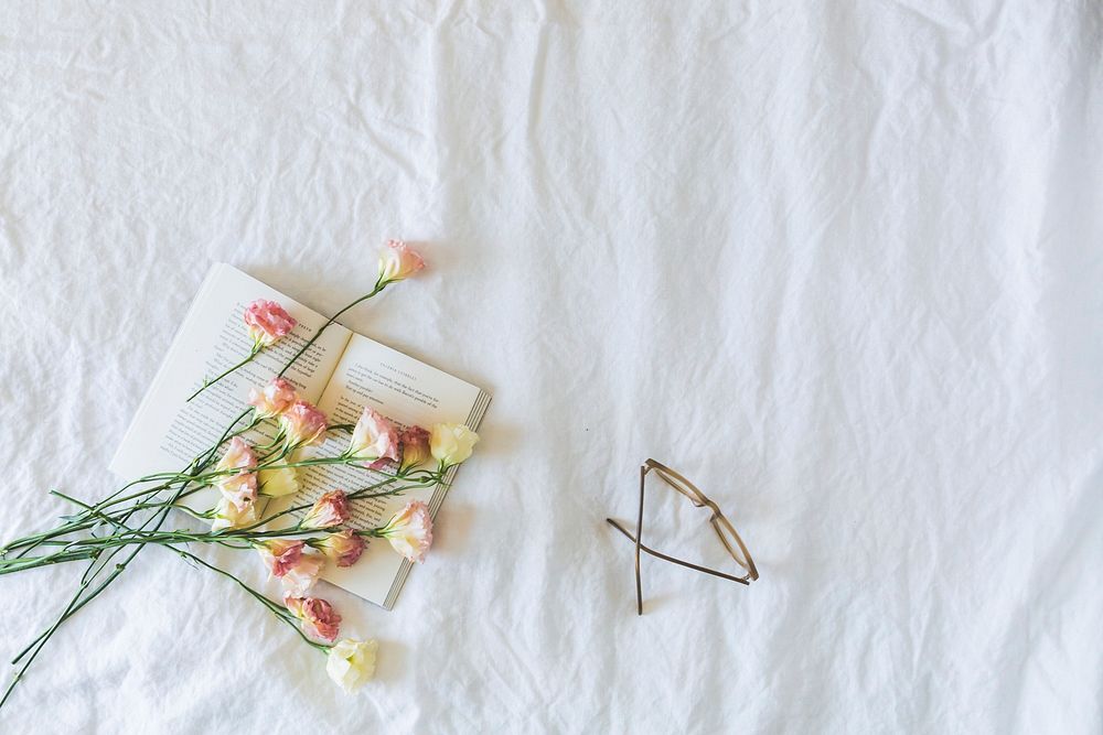 Pink and yellow flowers resting on a soft bed with glasses and a book.