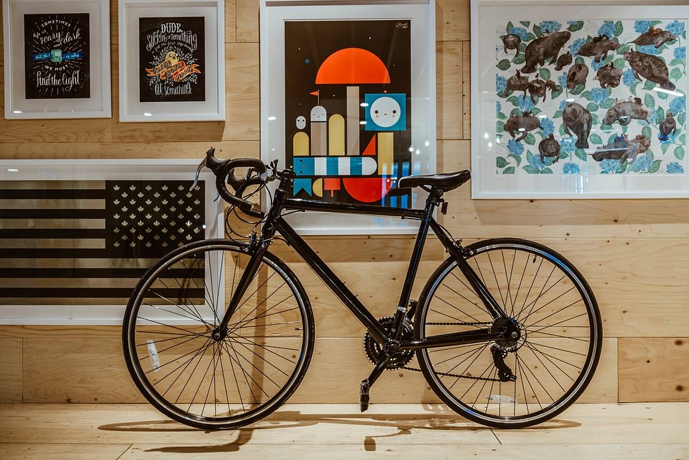 A sleek black road bike leans against a wall of art on a wooden indoor wall.