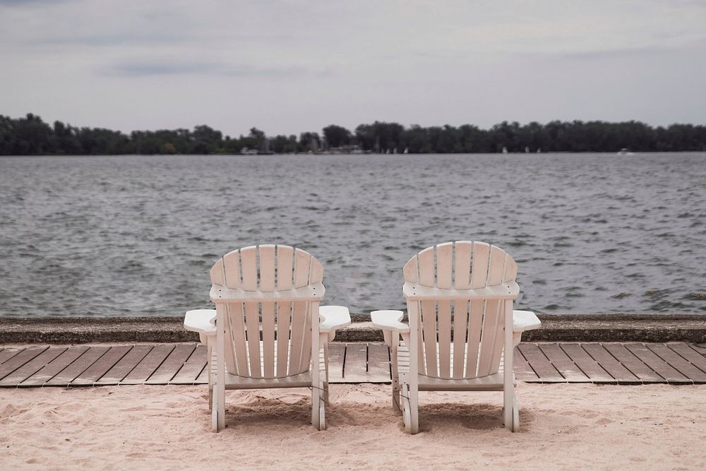 Two wooden beach chairs sit in the sand beside a boardwalk near a lake.