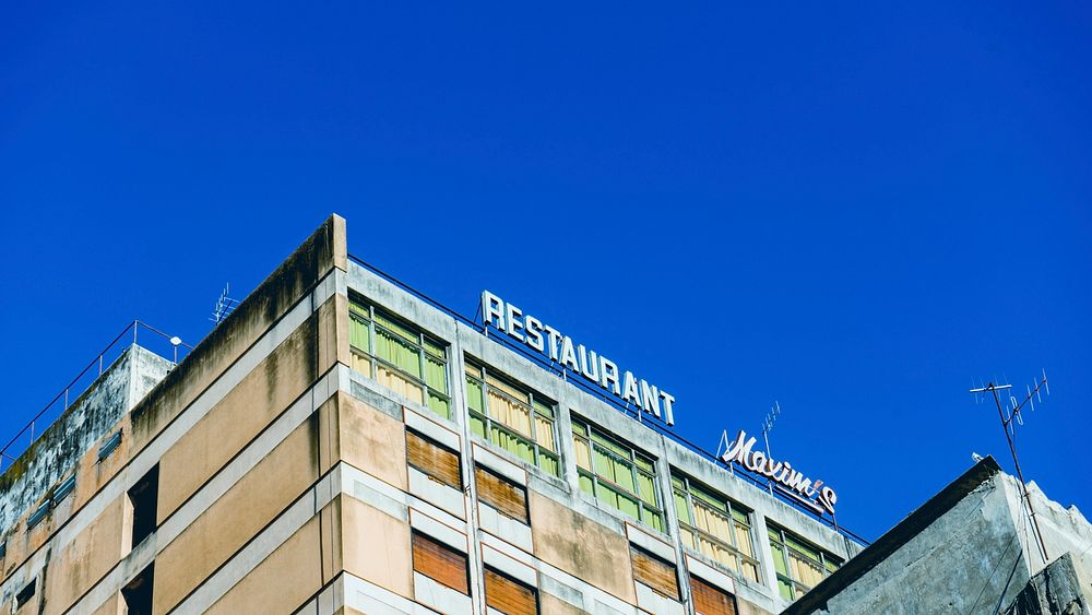 A sign for a restaurant stands on top of a party boarded up building with a clear sky above.