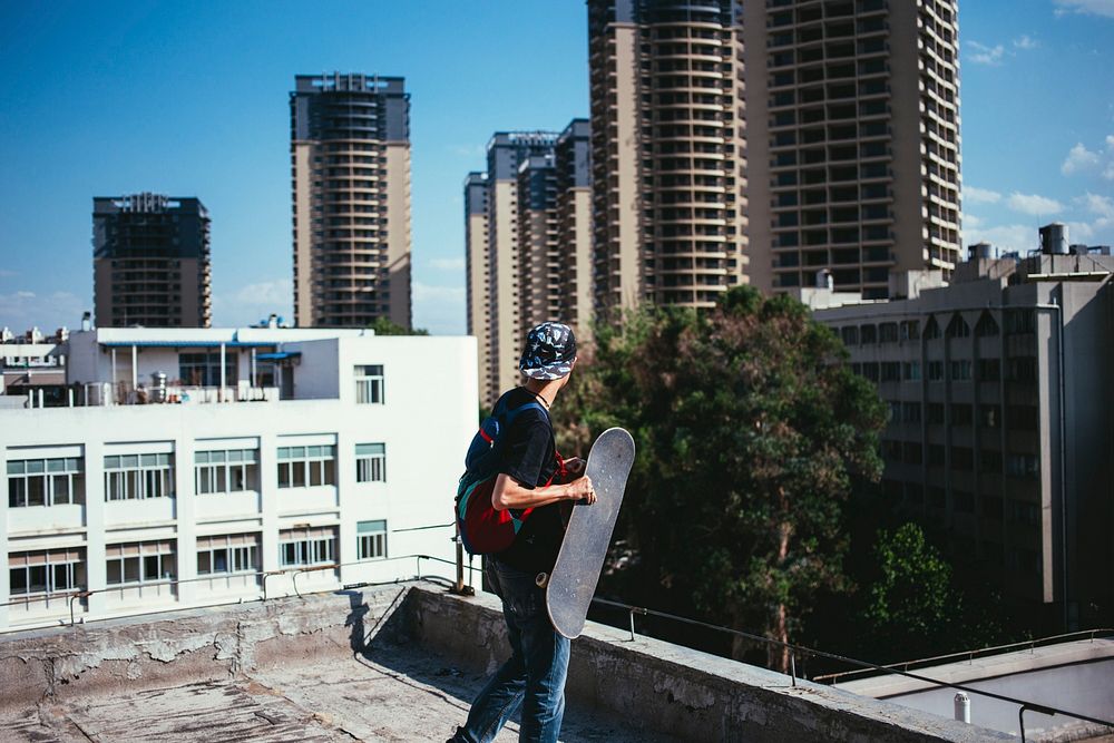 A person stands on a rooftop holding their skateboard and looking at the street below.