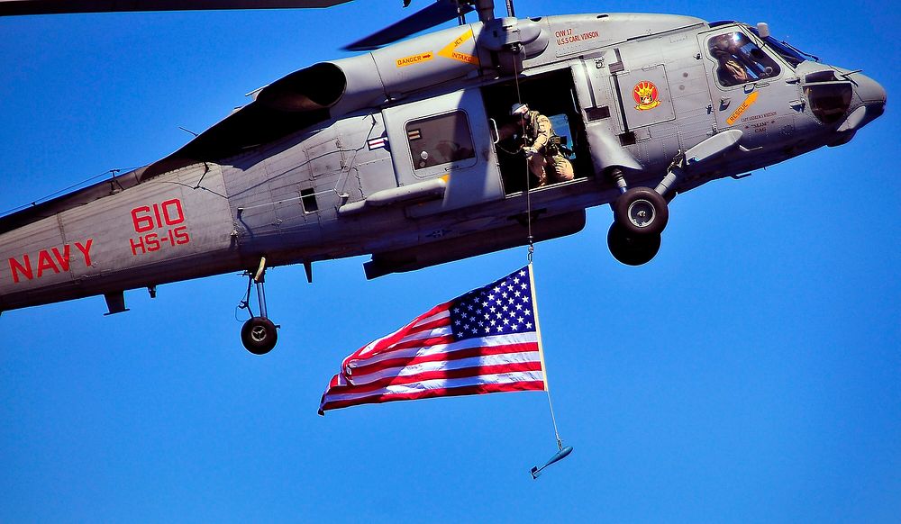 A U.S. Sailor from Explosive Ordnance Disposal Mobile Unit 3 suspends an American flag from an SH-60F Seahawk helicopter…