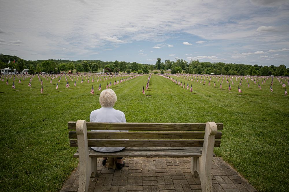 Elderly woman sits on a park bench remembering her husband, a Navy veteran, during a Memorial Day observance. Original…