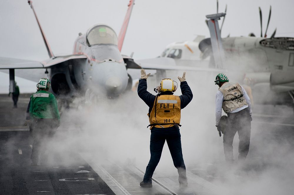 U.S. Navy Aviation Boatswain's Mate 2nd Class Tamara Sewell, center, directs an F/A-18C Hornet aircraft assigned to Strike…