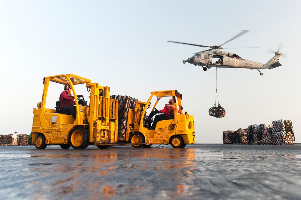 U.S. Sailors drive forklifts as an MH-60S Seahawk helicopter assigned to Helicopter Sea Combat Squadron (HSC) 22 delivers…