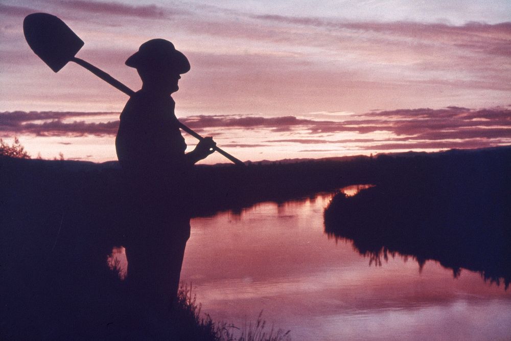 Silhouette of man holding shovel next to canal at sunset, scenic. Original public domain image from Flickr