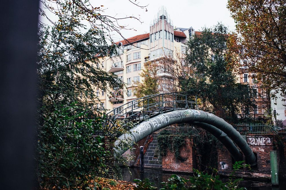 A curved metal pipeline over an urban river surrounded by buildings and trees