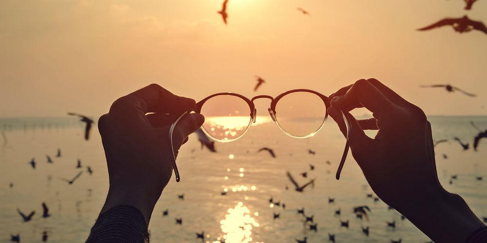 Hands holding out eyeglasses to sea and sunset