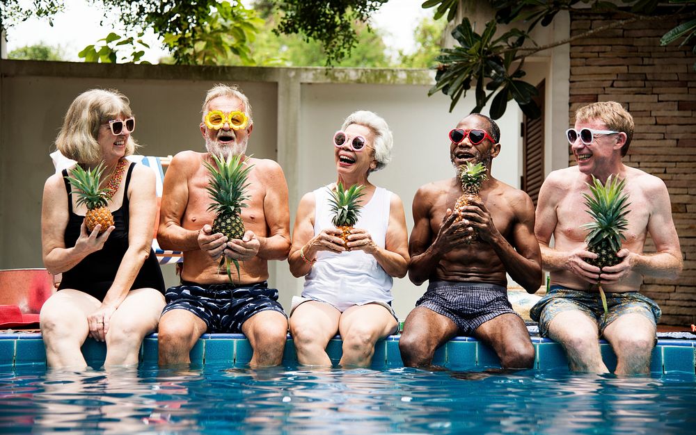 Group of diverse senior adults sitting at poolside holding pineapples together