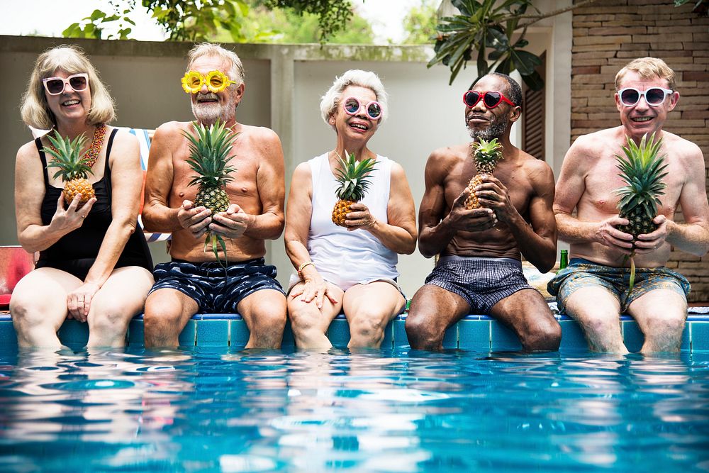 Group of diverse senior adults sitting at poolside holding pineapples together