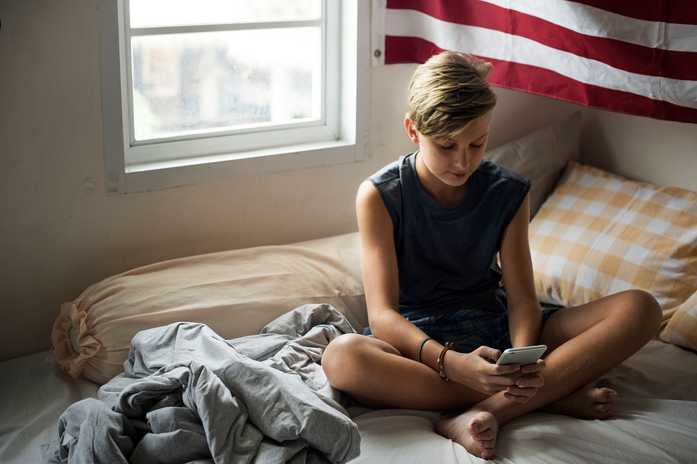 Young caucasian boy sitting using mobile phone on bed