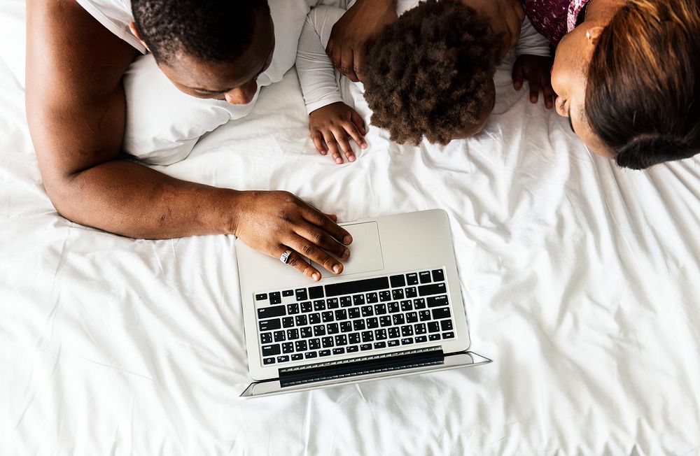 Black family lying on bed using computer laptop together in bedroom