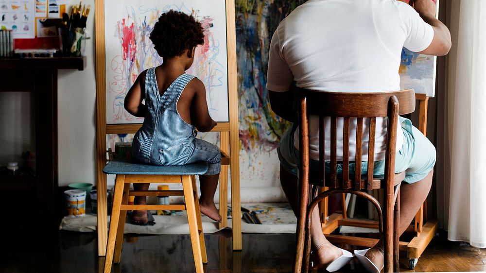 Black artist dad doing his art work with his child sitting nearby