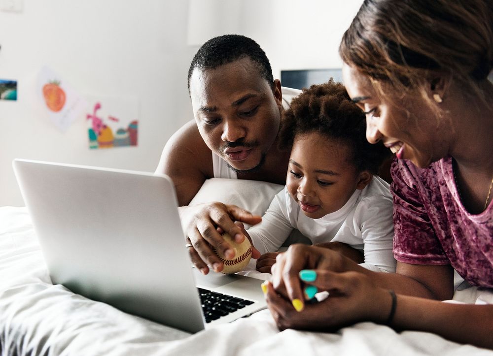 Black family lying on bed using computer laptop together in bedroom