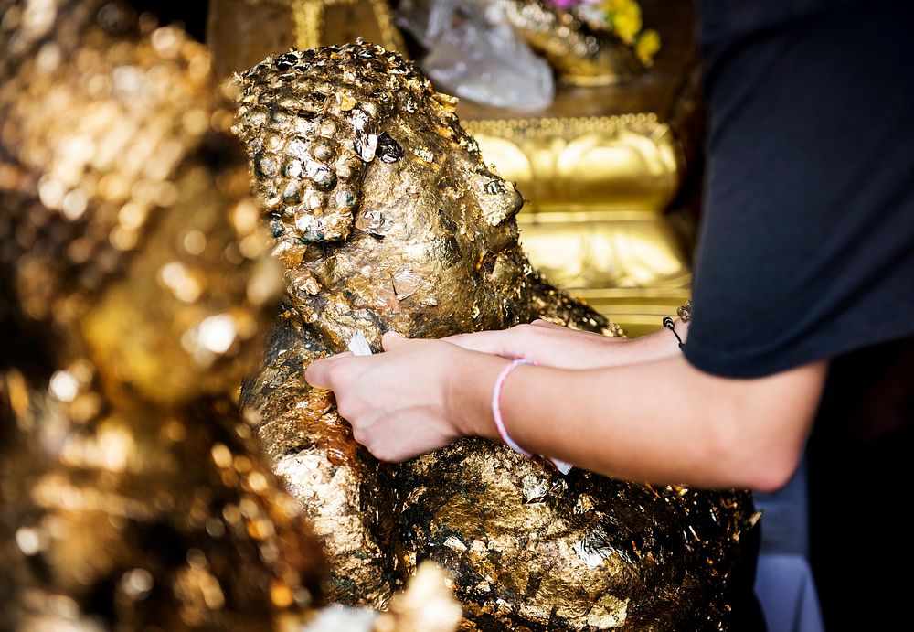 Closeup of hands gilding gold leaf sheet to Bhuddha statue in Thai temple