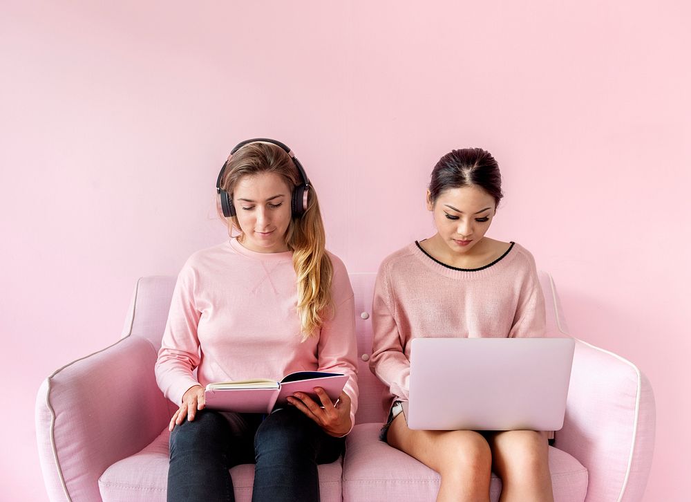 Women sitting together reading book and using computer laptop with pink background