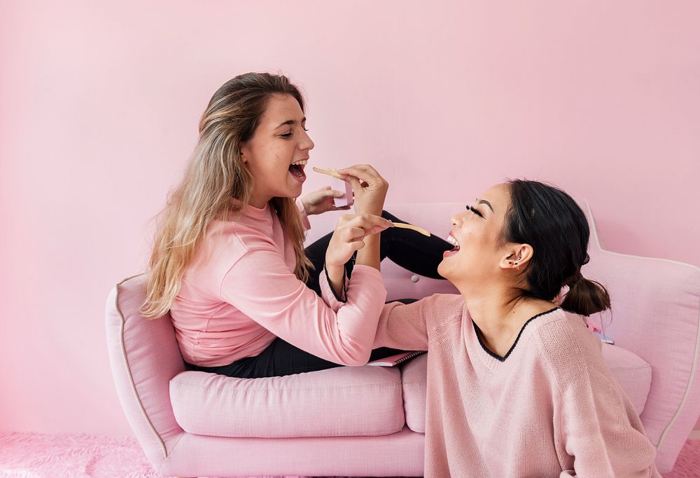 Women sitting feeding fries each other with pink background