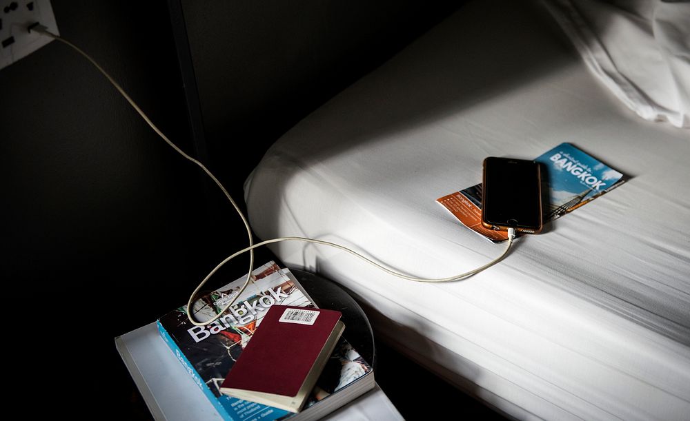 A mobile phone charging lying on the bed with a Bangkok, Thailand travel guide book and brochure