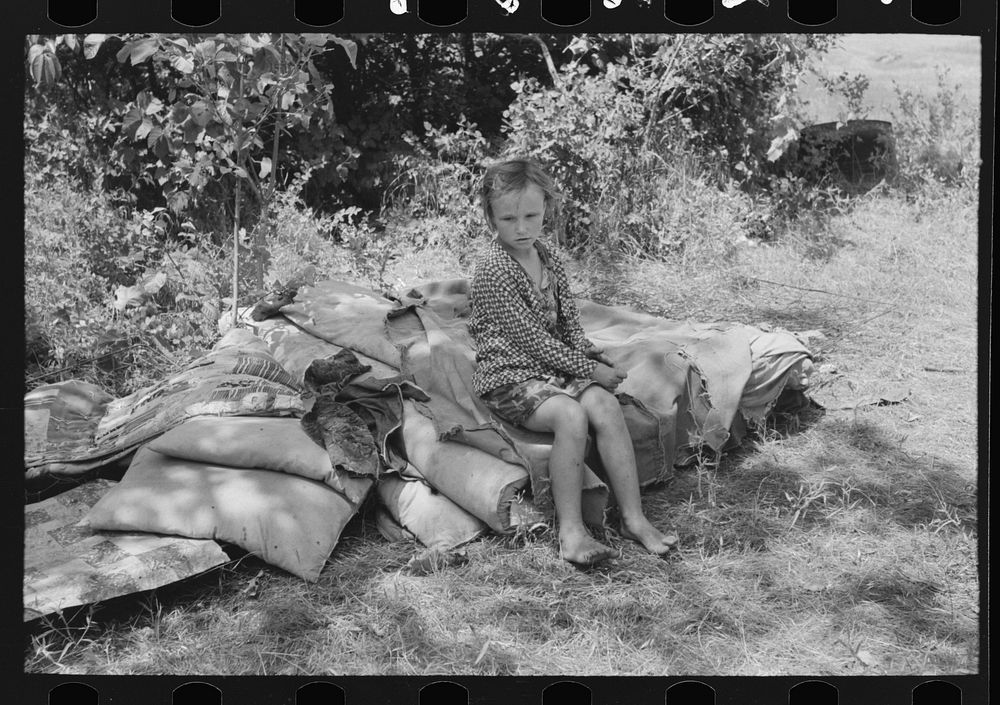 Child of family encamped by the roadside sitting on bedclothing.  There were no beds and no overhead covering near Spiro.…