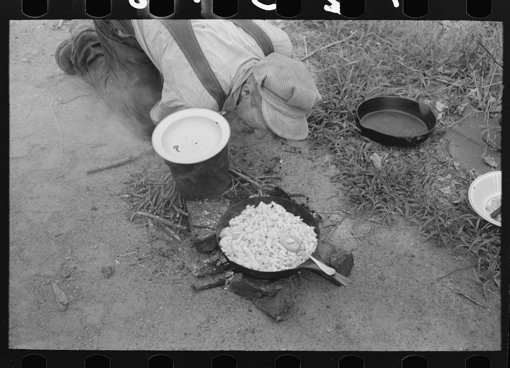 Nursing along the fire by blowing on it. Potatoes are being cooked in this pan. Migrant family en route to California near…