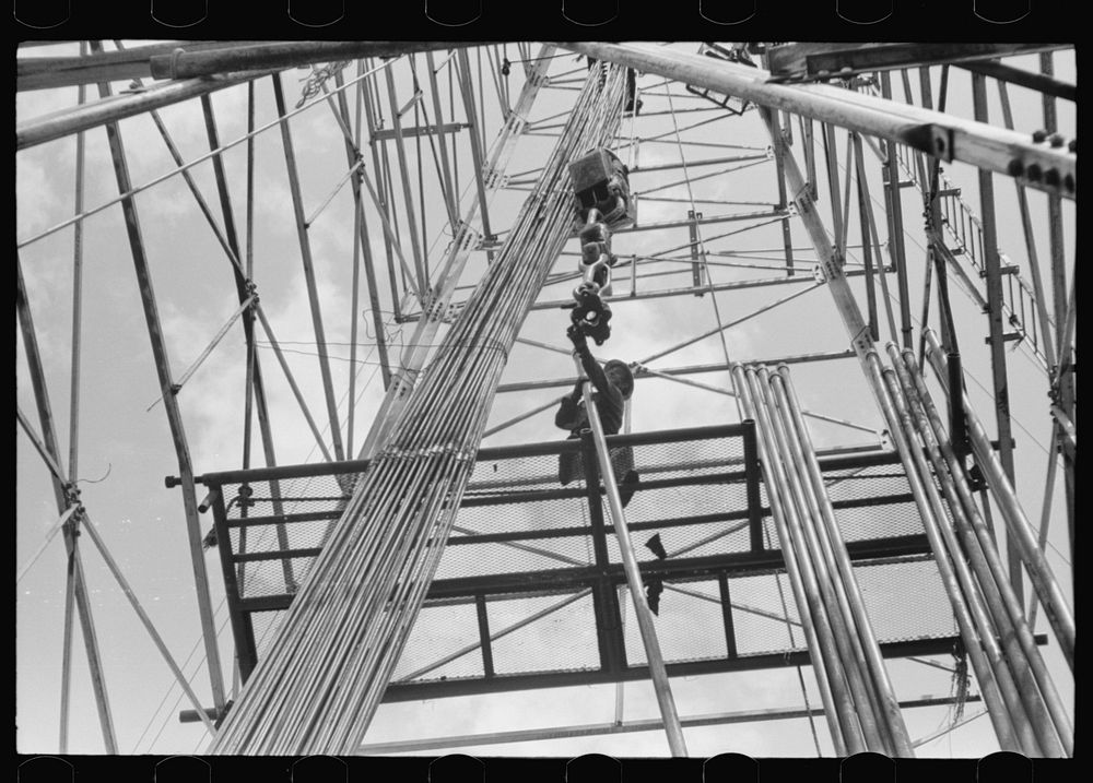 [Untitled photo, possibly related to: Looking up in an oil derrick. Roughneck inserting lengths of pipe into the elevator…