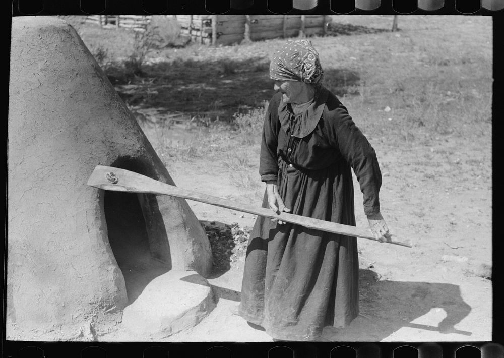 Spanish-American woman testing temperature of earthen oven by determining length of time required to scorch wool, Taos…