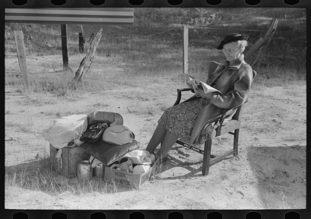Farmer's wife reading a magazine as she watches the family belongings which are parked beside the road while her husband has…