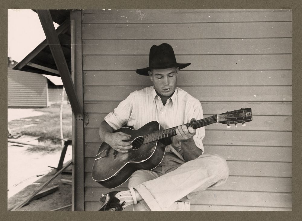 Migratory worker playing guitar on front porch of his metal shelter in the Agua Fria Labor Camp, Arizona by Russell Lee