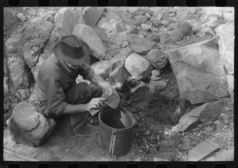 Prospector taking a sample of dirt from a creek bed. There is gold along this creek bed and he is trying, by taking samples…