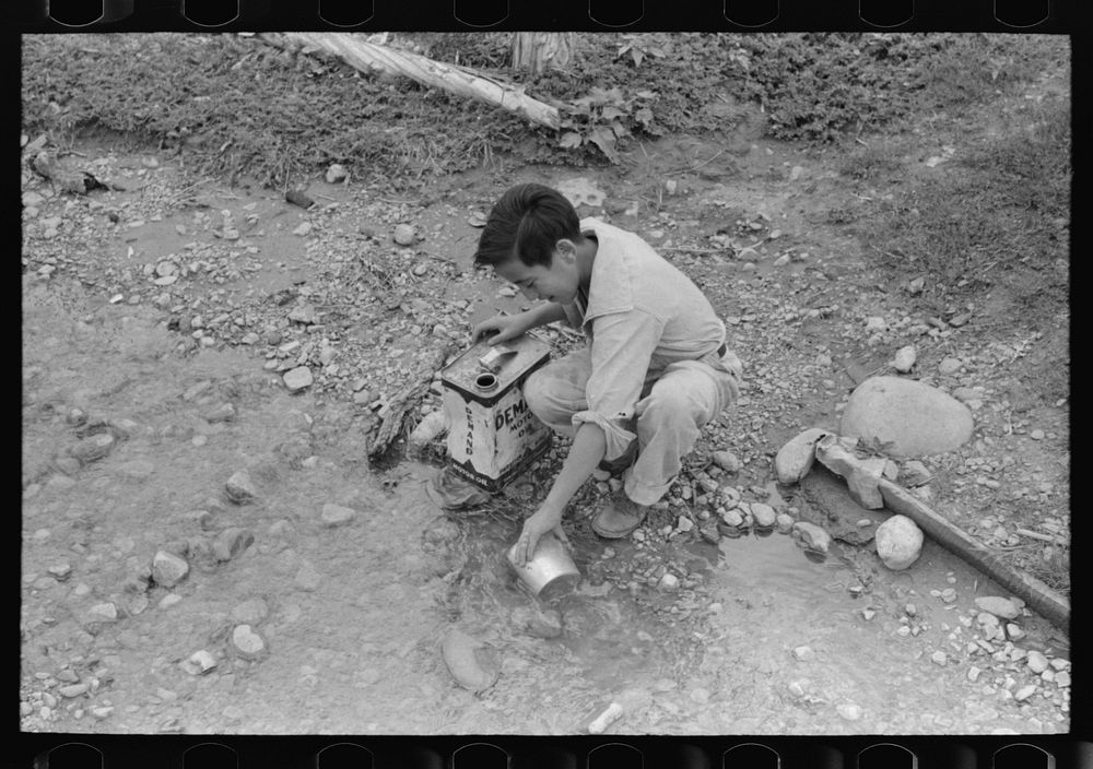 A Spanish-American boy dipping water from irrigating ditch. Water will be used in house for cooking, drinking, etc.…