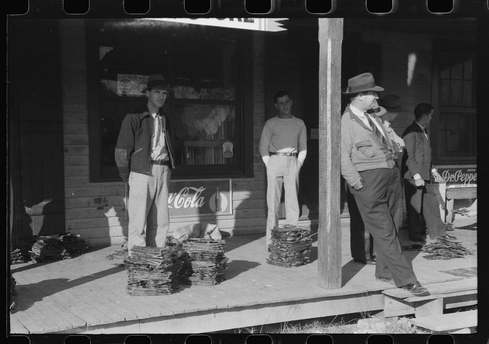 Spanish trappers and fur buyers waiting around while muskrats are being graded during auction sale on porch of community…