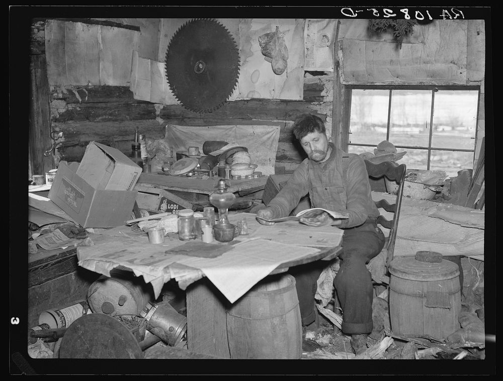 Buckboard Charlie reading the American Legion monthly in the living room of his shack near Iron River, Michigan by Russell…