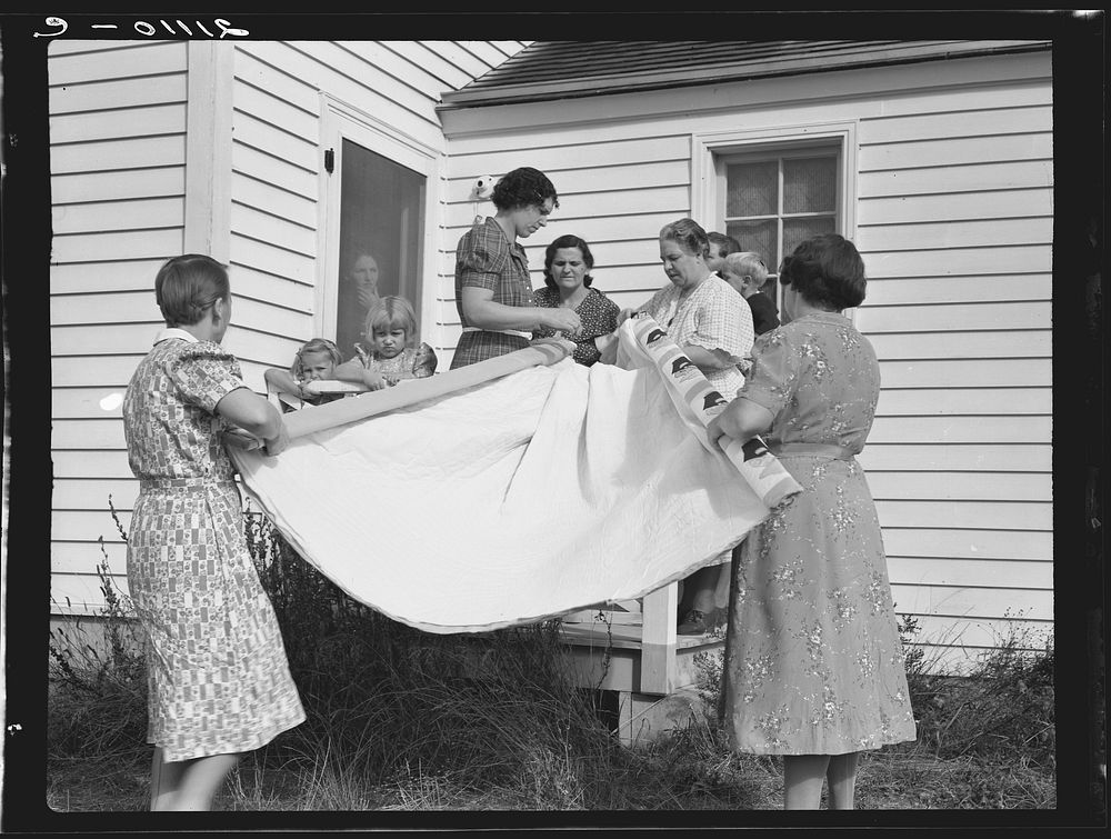 Farm women, members of the "Helping Hand" club, carefully roll up the quilt upon which they are working. Near West Carlton…