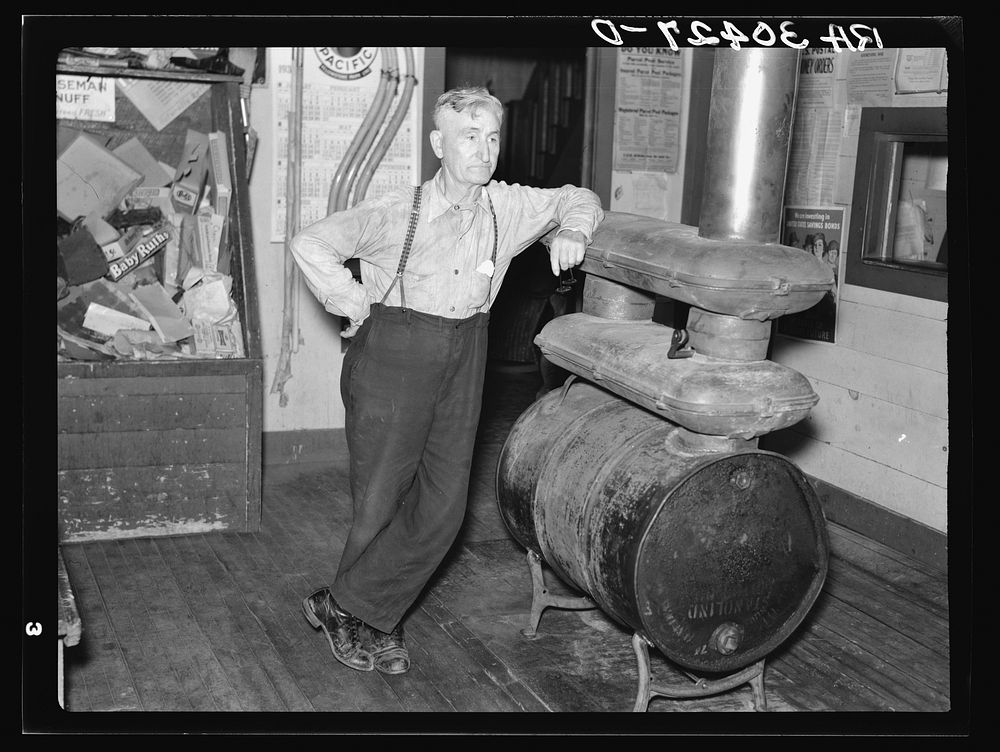 J. A. Fisher, storekeeper, postmaster and depot agent of Funkley, Minnesota, by stove of his own creation by Russell Lee