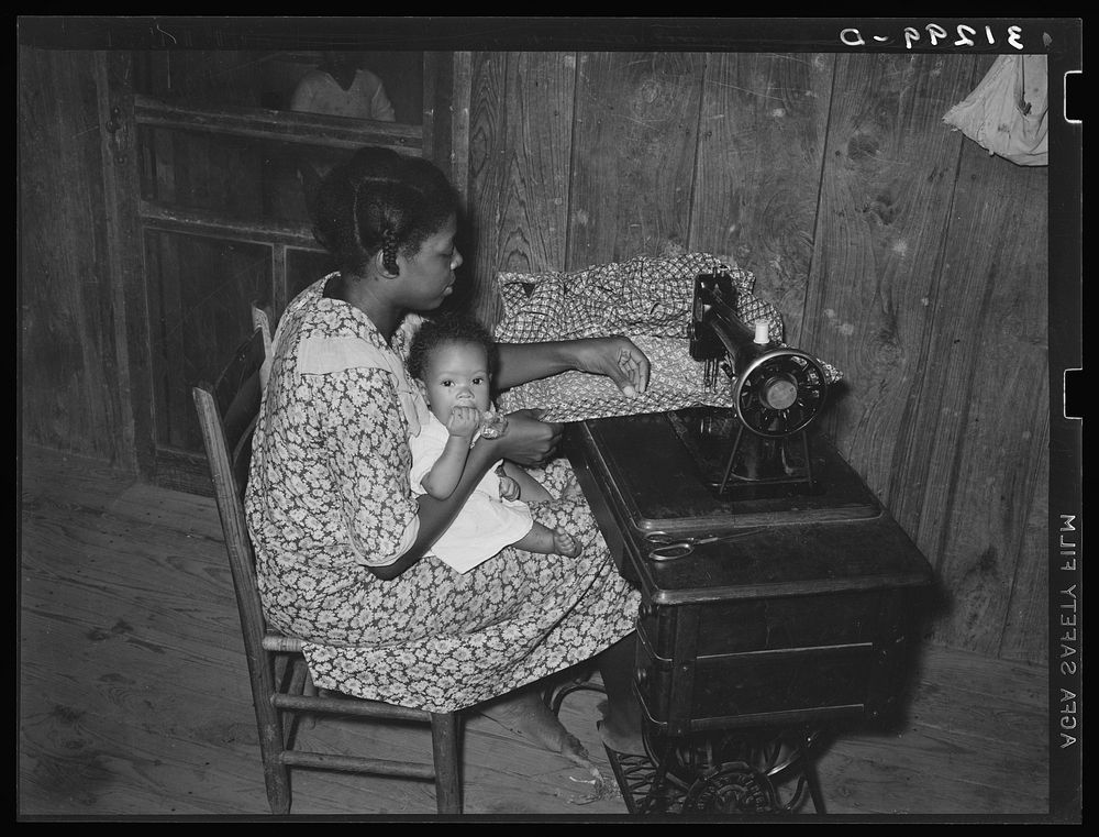Wife of sharecropper, with baby in her lap, at sewing machine. Family will work under tenant purchase program. Near…