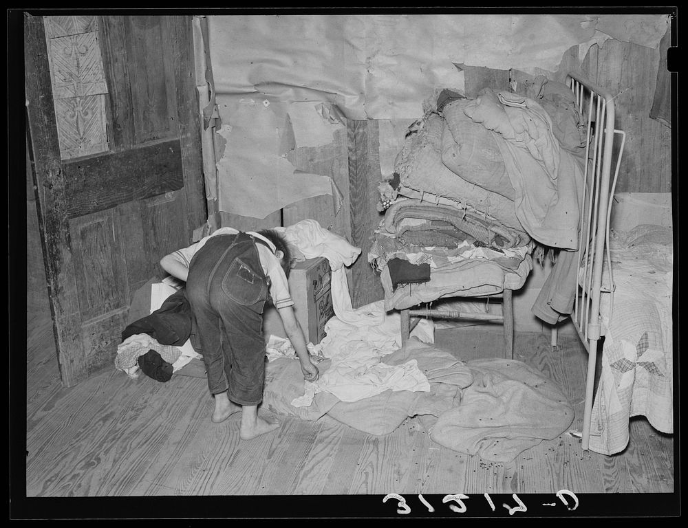 Southeast Missouri Farms. Child picking up clothes in corner of bedroom of shack home. Note lack of storage facilities by…