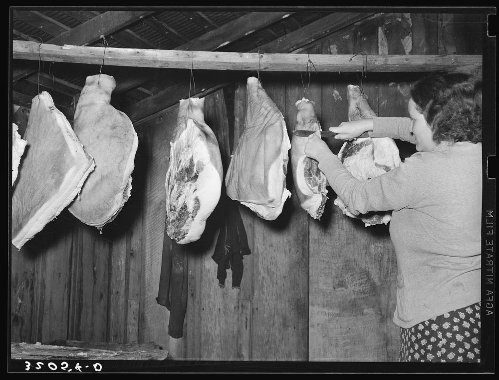 Wife of tenant farmer cutting piece of ham in smokehouse near Pace, Mississippi. Background photo for Sunflower Plantation…