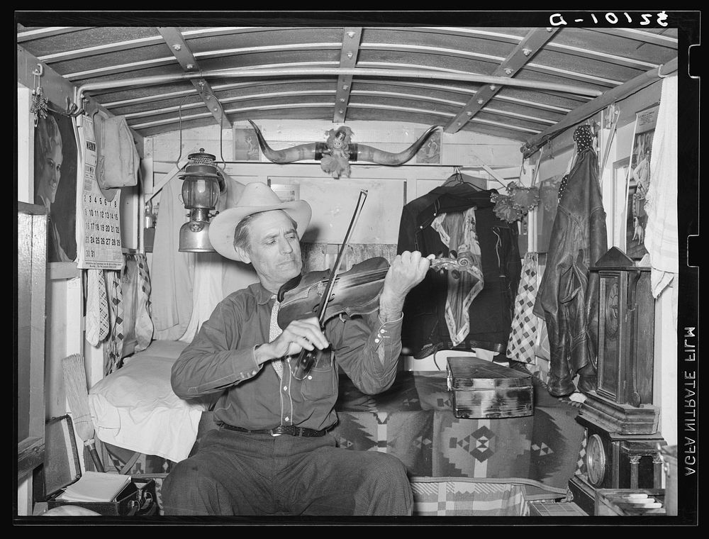 Mr. Bias playing the fiddle in his trailer home. He is a former cowboy who travels over the country. He has a small private…