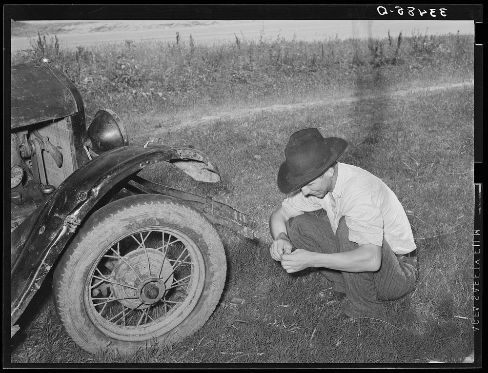 [Untitled photo, possibly related to: White migrant squatting down in front of his automobile while camped near Prague…